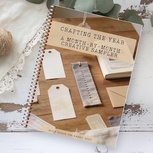 May include: A spiral-bound book titled "Crafting the Year: A Month-by-Month Creative Sampler" with blank tags, a fabric bookmark, and a ball of twine. The book is open, displaying various craft supplies on a wooden surface.