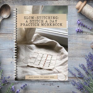 May include: A spiral-bound workbook titled "SLOW-STITCHING: A STITCH A DAY PRACTICE WORKBOOK" with a stitched fabric sample on the cover. The image includes a vintage spoon, spools of thread, lavender, and scissors, all arranged on a weathered wooden surface.