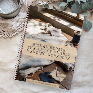 Op de afbeelding: Een spiraalgebonden werkboek getiteld "Ritual Repair: The Art of Visible Mending Workbook" met een beige omslag. Het boek ligt op een houten oppervlak met stofresten, een naald en draad. Een klos garen en een overhemd zijn ook zichtbaar.