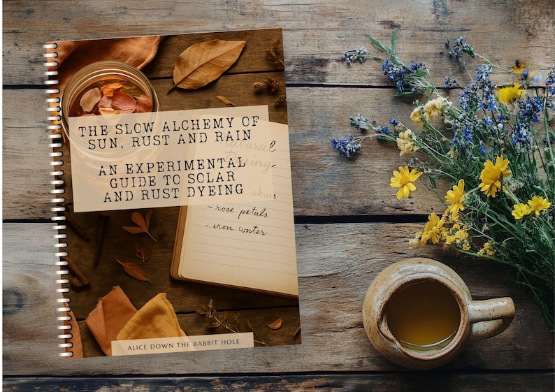 May include: A spiral-bound book titled The Slow Alchemy of Sun, Rust and Rain: An Experimental Guide to Solar and Rust Dyeing by Alice Down the Rabbit Hole, featuring a cover image of a jar, fabric scraps, and a notebook on a wooden surface.