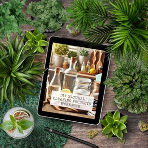 Op de afbeelding: Een digitale tablet met een werkboek met de titel "DIY Natural Cleaning Products Workbook" met een groene achtergrond en een foto van een keukenblad met schoonmaakmiddelen. De tablet is omringd door groene planten op een houten oppervlak.