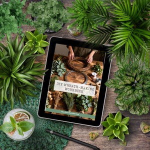 K&ouml;nnte beinhalten: Ein digitales Tablet mit dem Cover eines DIY-Kranz-Arbeitsbuchs auf einem Holztisch-Hintergrund, umgeben von verschiedenen gr&uuml;nen Topfpflanzen und einem schwarzen Eingabestift. Der Bildschirm zeigt eine Szene zur Kranzherstellung.