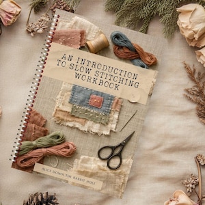 Op de afbeelding: Een spiraalgebonden werkboek met de titel AN INTRODUCTION TO SLOW STITCHING WORKBOOK door ALICE DOWN THE RABBIT HOLE. De kaft bevat stoffen lapjes, borduurgaren in groen, roze en blauw, een houten klosje, een naald en een zwarte schaar.