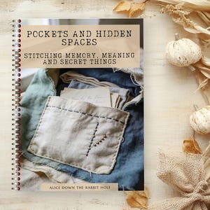 May include: A spiral-bound book titled "Pockets and Hidden Spaces" with the subtitle "Stitching Memory, Meaning and Secret Things." The cover features a close-up of a stitched pocket and fabric. Decorative pumpkins and foliage are in the background.