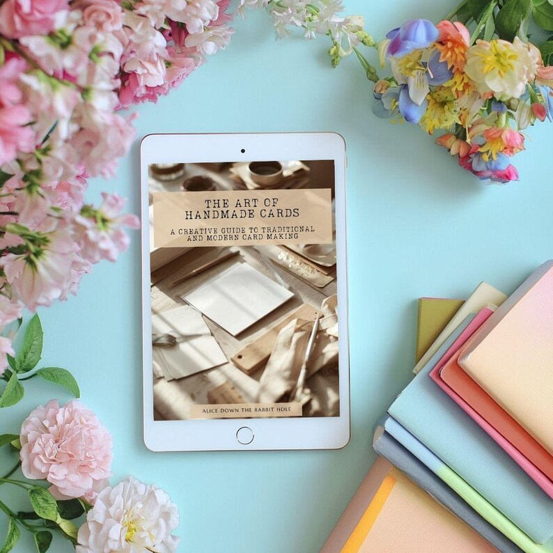 Pu&ograve; includere: Un tablet bianco che mostra la copertina di un libro intitolata The Art of Handmade Cards: A Creative Guide to Traditional and Modern Card Making di Alice Down the Rabbit Hole, con articoli di cancelleria su sfondo azzurro.