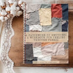 Pode incluir: Um caderno espiralado intitulado "A Patchwork of Emotions: A Workbook for Feeling Through Fabric" com uma capa de patchwork multicolorida. O livro está sobre uma superfície de madeira, acompanhado por ramos de algodão e renda.