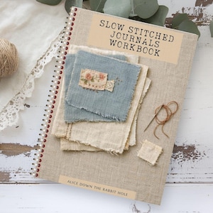 May include: A spiral-bound workbook titled SLOW STITCHED JOURNALS WORKBOOK and ALICE DOWN THE RABBIT HOLE. The beige linen cover features layered fabric swatches in blue and cream, a small floral patch, a sewing needle, and brown thread.