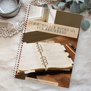 Puede incluir: Un cuaderno con espiral titulado "Commonplace Books 101 for Beginners" está abierto, mostrando notas manuscritas. Un bolígrafo y un lápiz descansan sobre la superficie de madera. El libro está sobre una superficie de tono neutro con un borde de encaje.