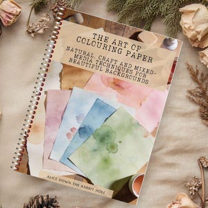 May include: A spiral-bound book titled "The Art of Colouring Paper" with watercolor paper samples in pink, blue, and green. The book is surrounded by dried flowers, pinecones, and greenery, creating a rustic aesthetic.