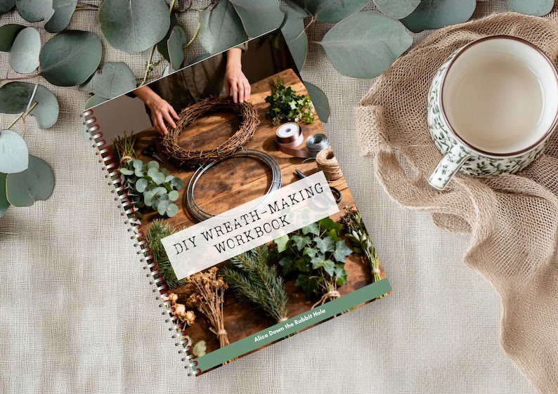 K&ouml;nnte beinhalten: Ein spiralgebundenes Arbeitsbuch mit dem Titel DIY WREATH-MAKING WORKBOOK, dessen Einband eine Holzoberfl&auml;che, verschiedene Gr&uuml;npflanzen, Kranzrahmen und B&auml;nder zeigt. Am unteren Rand steht der Text Alice Down the Rabbit Hole.