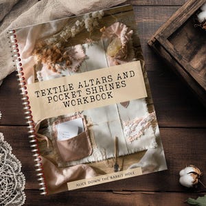 May include: A spiral-bound workbook titled "TEXTILE ALTARS AND POCKET SHRINES WORKBOOK" with a beige cover, featuring fabric and floral elements. The book lies on a dark wooden surface, with cotton bolls in a wooden crate nearby. The text "ALICE DOWN THE RABBIT HOLE" is also visible.