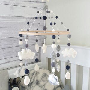 May include: A baby mobile with a wooden ring and hanging felt shapes. The mobile features white, gray, and navy blue felt balls, clouds, stars, and teardrop shapes. The mobile hangs above a crib with pillows and a stuffed animal.