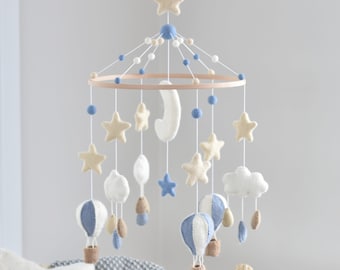 Könnte beinhalten: Ein Baby-Mobile mit einem Holzring, blauen und weißen Filzwolken, Sternen, Heißluftballons und einem Halbmond. Das Mobile ist mit blauen und weißen Pompons verziert.