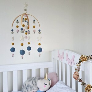 May include: A wooden baby mobile with felt stars, balls, and beads in shades of gray, pink, and gold. The mobile is hanging above a white crib with a stuffed unicorn toy.