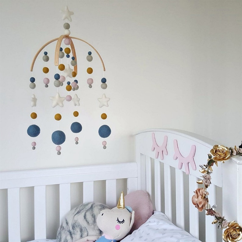May include: A wooden baby mobile with felt stars, balls, and beads in shades of gray, pink, and gold. The mobile is hanging above a white crib with a stuffed unicorn toy.