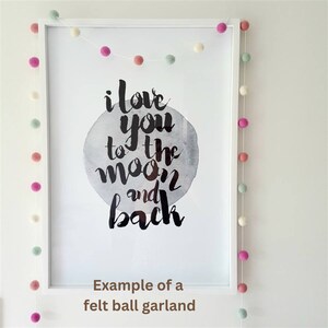 May include: A white frame with a print that says "i love you to the moon and back" with a gray watercolor moon. A felt ball garland with white, pink, green, and cream balls hangs around the frame.