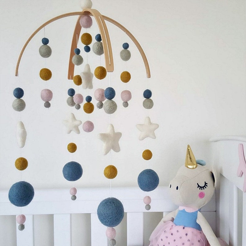May include: A wooden baby mobile with felt balls in shades of blue, pink, gray, and yellow. The mobile also features white felt stars.