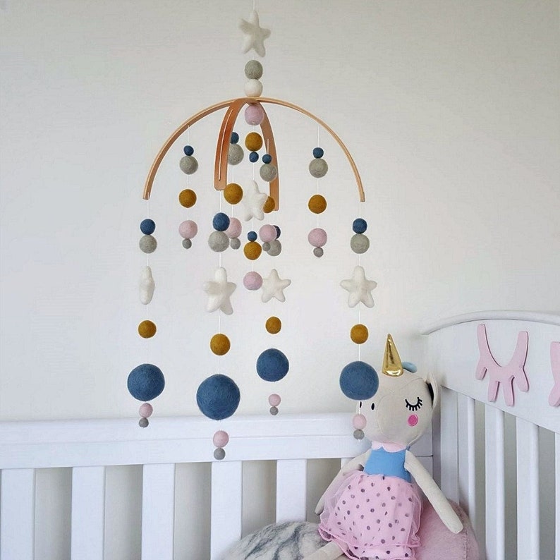 May include: A wooden baby mobile with felt stars, balls, and beads in shades of blue, gray, pink, and yellow. The mobile is hanging above a white crib with a stuffed unicorn toy.