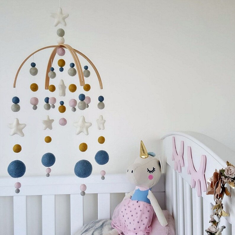 May include: A wooden baby mobile with felt stars, balls, and circles in shades of blue, gray, pink, and yellow. The mobile is hanging above a white crib with a pink unicorn stuffed animal.