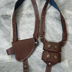 Puede incluir: Un holster de cuero marrón con dos fundas para cargadores. El holster tiene una correa para el hombro y está diseñado para llevarse en el cuerpo.