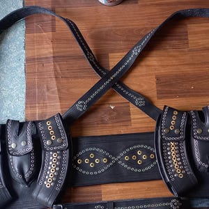 以下が含まれることがあります： 複数のポーチとストラップが付いた黒革のチェストハーネス。ハーネスは金色のリベットと装飾的な切り抜きが特徴です。デザインには、コスプレやコスチュームに適したショルダーストラップとウエストベルトが含まれています。