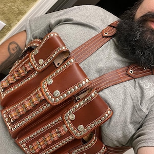 Può includere: Una cintura portautensili in pelle marrone con più tasche e borchie argentate. La cintura ha un design intrecciato sul davanti ed è indossata sopra una camicia grigia.