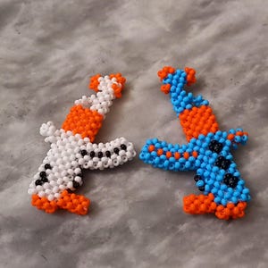 May include: Two small beaded figures, one white and one blue, with orange accents. The figures are shaped like guns and are made of small beads.