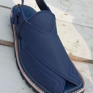 May include: A pair of dark blue leather sandals with a strap and buckle. The sandals have a decorative stitching pattern and a white trim around the sole.