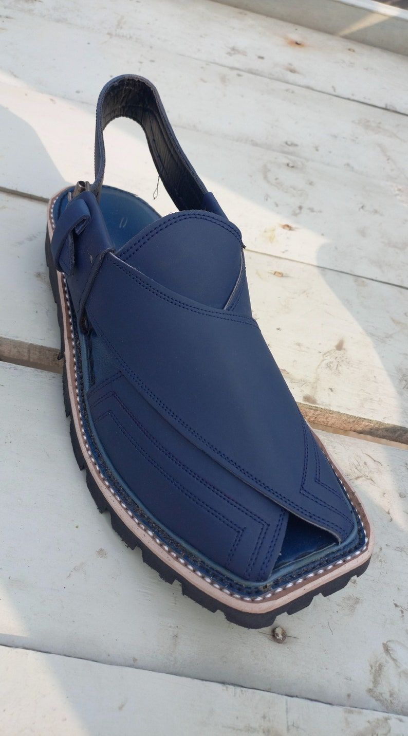 May include: A pair of dark blue leather sandals with a strap and buckle. The sandals have a decorative stitching pattern and a white trim around the sole.