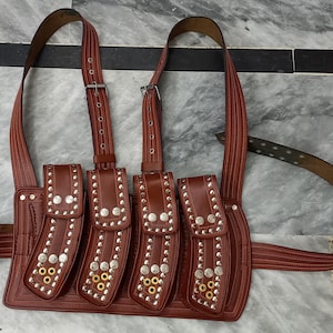 May include: A brown leather belt with multiple pouches and silver studs. The belt has a wide strap and is adjustable.
