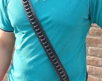 Peut inclure: Un holster en cuir noir avec une bandoulière et une boucle. La sangle comporte une série de boucles décoratives. Le holster est porté par une personne portant un polo bleu avec le texte "Leisure Club" sur la poitrine.