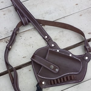 May include: A brown leather shoulder holster with white stitching and a silver buckle. The holster has a magazine pouch and a flap to secure a firearm.