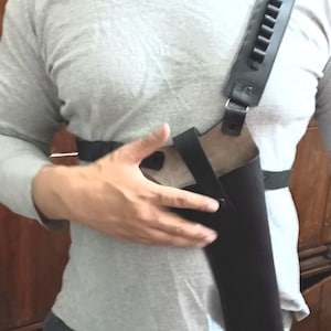 May include: Black leather shoulder holster with a vertical gun pocket and a strap with bullet loops. The holster is worn across the chest and secured with a waist strap. The design is for carrying a firearm.