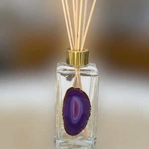 Puede incluir: Un frasco difusor de vidrio transparente con una tapa dorada y una rodaja de ágata púrpura. El frasco contiene varillas de madera para la difusión de la fragancia. La rodaja de ágata tiene un patrón concéntrico púrpura y blanco.