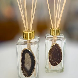 May include: Two clear glass reed diffusers with gold-colored collars and decorative agate slices. Each diffuser has a bundle of light-colored reeds. The agate slices have brown, gray, and white bands. The diffusers are square-shaped.