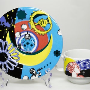 Op de afbeelding: Een keramieken schoteltje en kopje met een levendig, meerkleurig grafisch ontwerp. Het schoteltje toont een clown, een gele maansikkel, een paarse touwknoop en muzieknoten. Het kopje is voorzien van een klassieke buste en abstracte vormen.