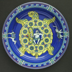 Op de afbeelding: Een decoratieve bord met een blauwe achtergrond en een geel schildpadontwerp. De schildpadschild heeft een centrale figuur en bloemaccenten. De rand van het bord heeft een geometrisch patroon en bevat kleine dierfiguren.