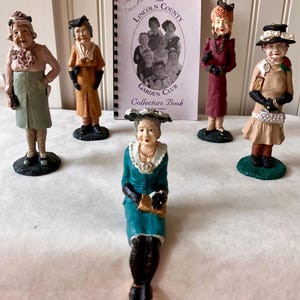 May include: A collection of vintage figurines depicting elderly women in various outfits. The figurines are painted with detailed features and stand on small bases. A book titled "The Lincoln County Garden Club Collectors Book" is also present.
