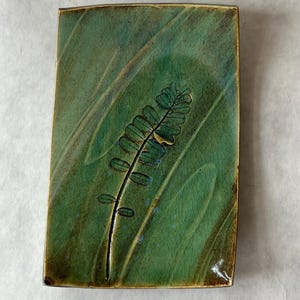 May include: Rectangular ceramic tray with a green glaze and a fern design. The tray has a dark brown border and a crackle finish. The fern is black and has small, oval-shaped leaves. The tray is approximately 6 inches long and 4 inches wide.