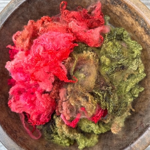 May include: A collection of loose, unspun wool fibres in vibrant shades of bright red, deep pink, and olive green, contained within a dark, circular wooden bowl. The fibres have a soft, curly, and textured appearance, showing natural variations.