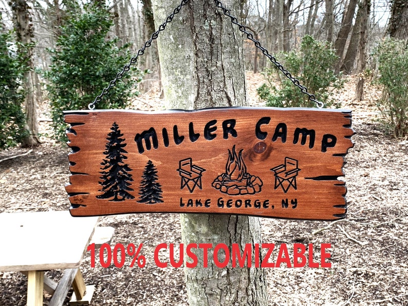 May include: A wooden sign with the text "MILLER CAMP" and "LAKE GEORGE, NY" in black lettering. The sign also features a campfire, two camping chairs, and two pine trees. The sign is hanging from a chain.