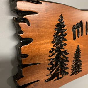 May include: A wooden sign with a rustic, natural edge. The sign is stained a rich brown color and features a black silhouette of two pine trees, a camping chair, and the text "Miller Lake Glen".
