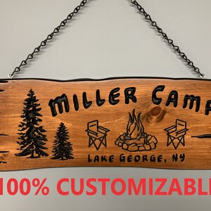 May include: A wooden sign with the text "MILLER CAMP" in black lettering. The sign also features two stylized trees, a campfire, and two camping chairs. The text "LAKE GEORGE, NY" is also included on the sign.