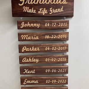 May include: A wooden sign with the text "Grandkids Make Life Grand" at the top. Below the text are seven wooden plaques with the names of grandkids and their birth dates: Johnny 04-12-2015, Maria 10-22-2016, Parker 04-02-2017, Ashley 01-02-2019, Kent 09-06-2021, Emma 05-09-2022, and Maggie 02-22-2023.