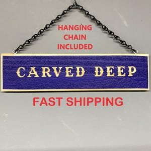 May include: A wooden sign with a blue background and gold lettering that reads "CARVED DEEP". The sign is hanging from a black chain and has the text "HANGING CHAIN INCLUDED" and "FAST SHIPPING" printed below.