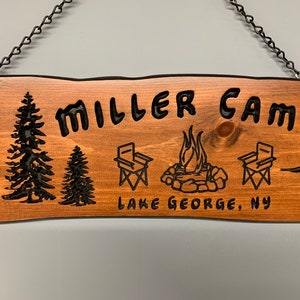 May include: A wooden sign with the text "Miller Camp" and "Lake George, NY". The sign is decorated with a campfire, two camping chairs, and two pine trees. The sign is hanging from a chain.