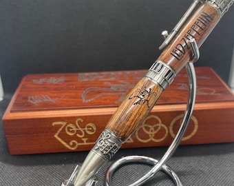 May include: A wooden pen with metal accents, featuring the engraved text "LED ZEPPELIN". The pen has a detailed design with a music-themed band and a stylized insect. It rests on a silver pen stand, with a wooden box in the background.