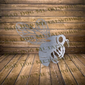 May include: A metal cutout of a bee with detailed wings and a textured body. The bee is in flight and has a large, round eye. The cutout is on a wooden background.
