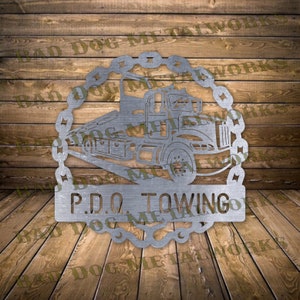 Könnte beinhalten: Ein silbernes Metallschild mit einem Abschleppwagen-Design in einem Kettenmuster. Der Schriftzug lautet "P.D.Q. TOWING". Das Schild ist auf einem Holzgrund montiert, wahrscheinlich für ein Abschleppunternehmen.