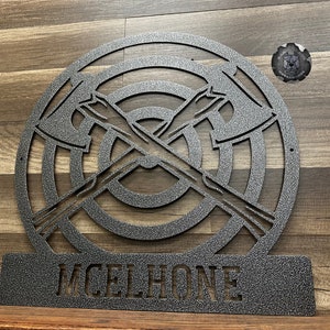 May include: A metal wall decor featuring a stylized axe throwing target with two crossed axes. The target has concentric circles and the name "McElhone" is displayed on a separate metal plaque below.